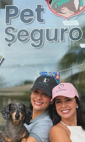 Duas mulheres com bonés da Pet Seguro e um dachshund na frente da fachada da loja